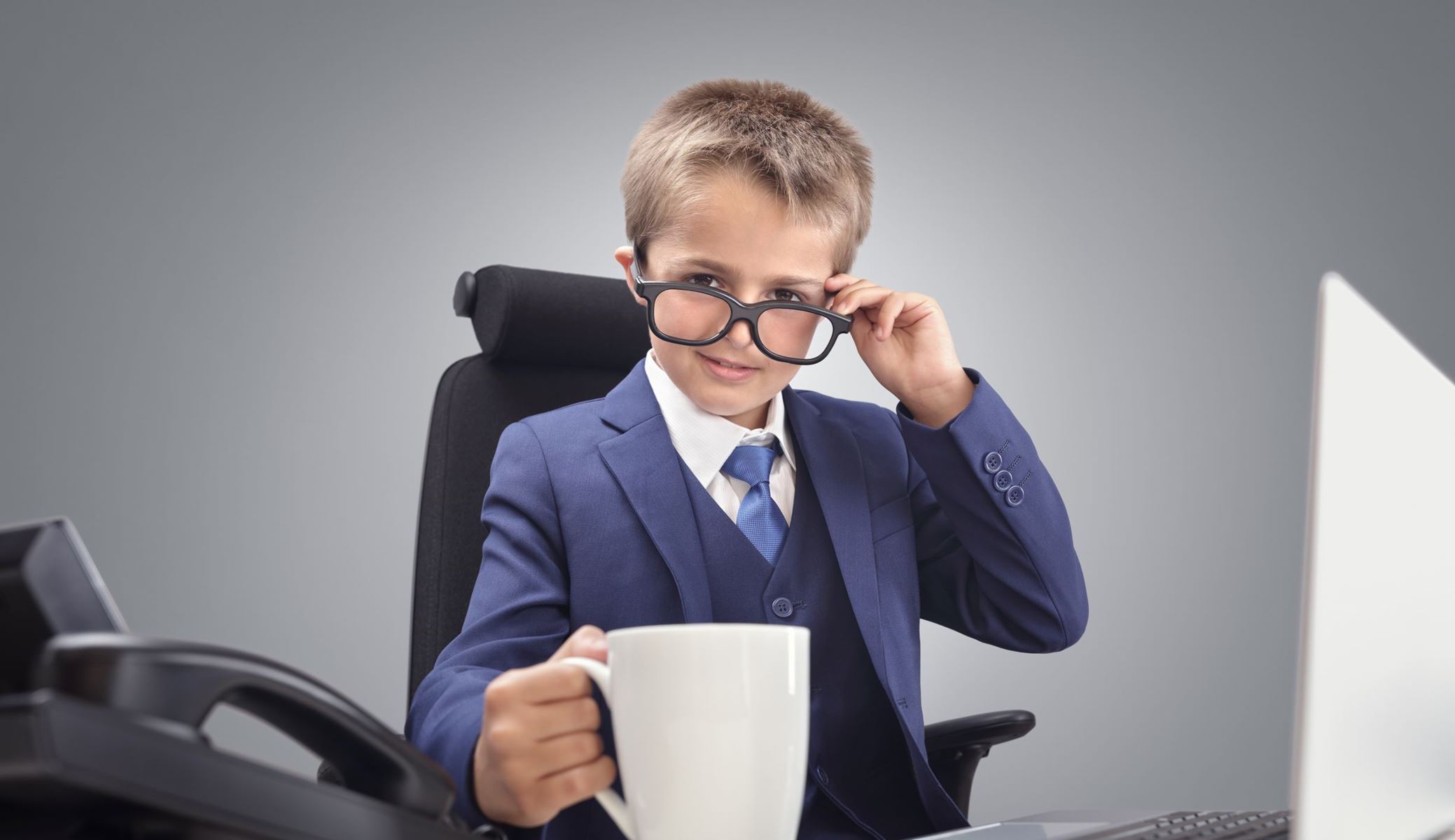 Kid dressed in a suit and tie, sitting at a computer with a coffee mug